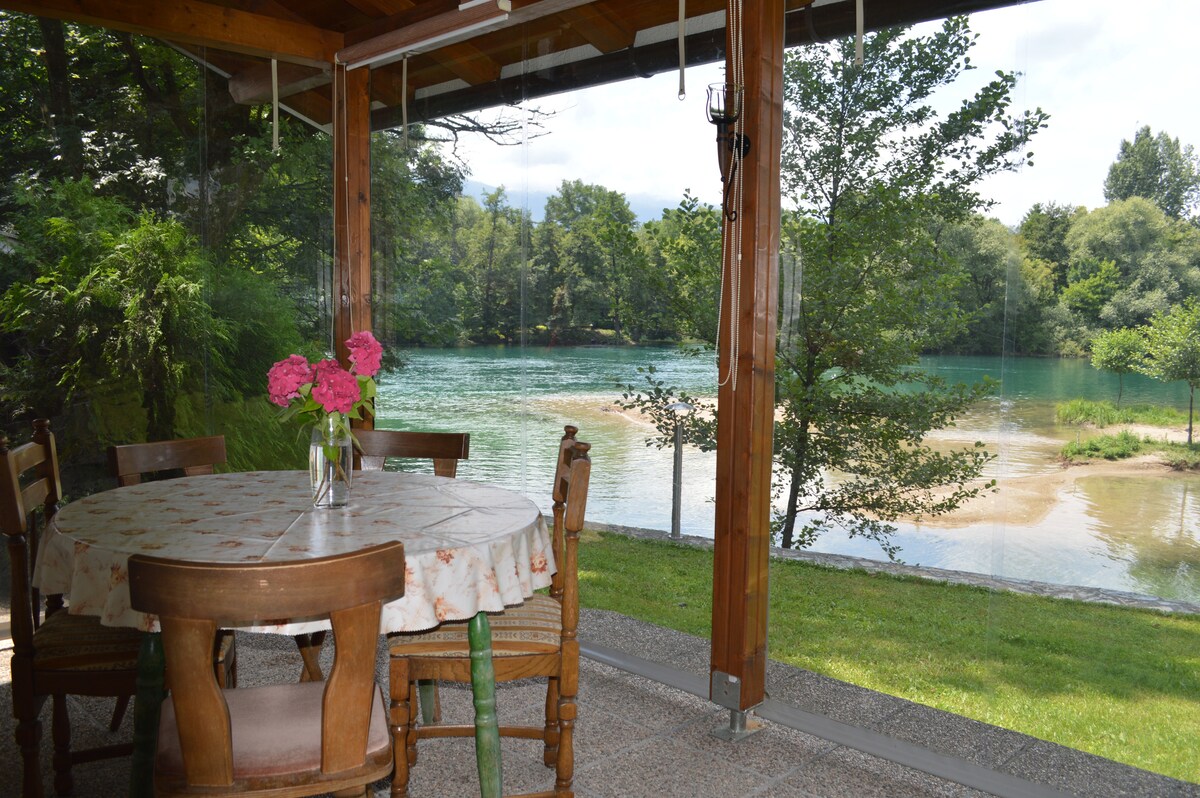 A covered outdoor dining area features a round table set for six, with a bouquet of pink flowers at its center. Glass panels provide unobstructed views of the river and surrounding greenery, creating a seamless connection between the interior space and the natural environment.