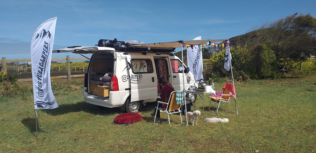 A mini camper is parked on grassy ground, featuring an extended awning that provides shade. Nearby, a table is set with colorful chairs, and a small dog is present. Flags representing the rental company are displayed, indicating a welcoming outdoor space for relaxation.