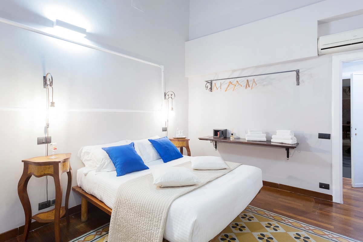 A spacious guest room is furnished with a large bed adorned with two blue pillows. A wooden shelf holds neatly arranged white towels. The room features warm wooden flooring and soft lighting, complemented by air conditioning and tasteful wall decor.