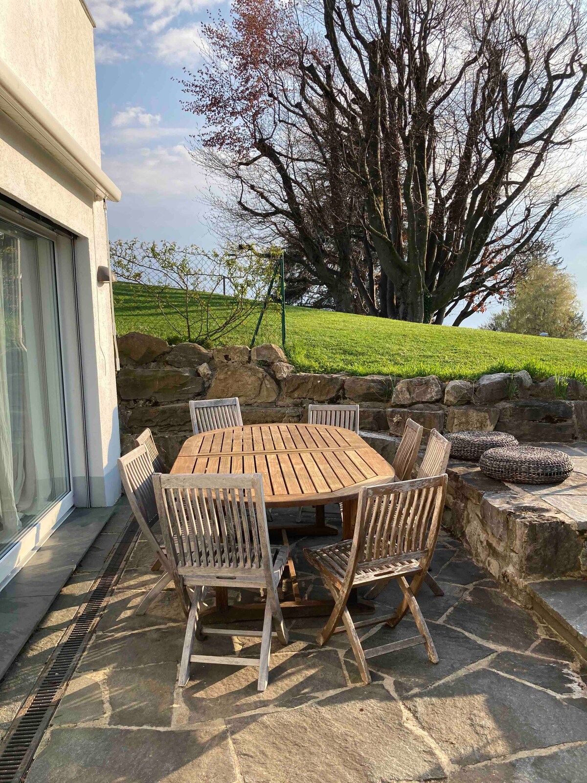 An outdoor terrace is furnished with a wooden dining table surrounded by six matching chairs. The stone-paved area is complemented by a natural stone wall and offers a view of a lush green lawn and a large tree in the background.
