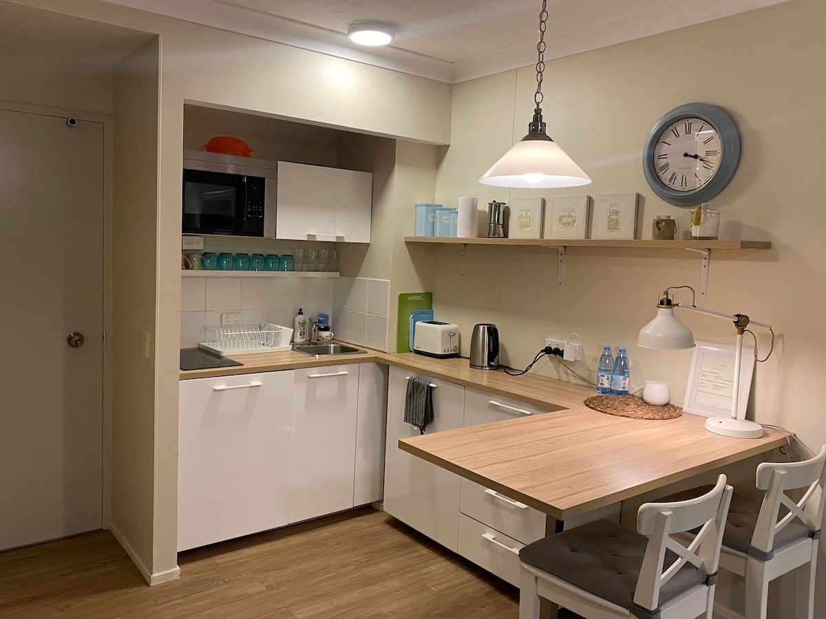 A modern kitchen area features white cabinetry and a wooden countertop. Essential appliances are neatly arranged, including a microwave, kettle, and a compact refrigerator. A dining table with two chairs sits against the wall, complemented by decorative items and a wall clock.