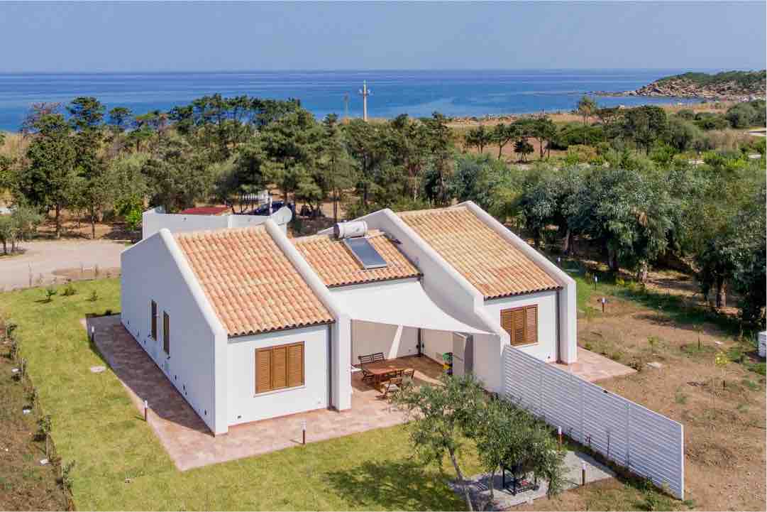 The exterior of a modern white house is viewed from above, showcasing a terracotta-tiled roof. Surrounding greenery and mature trees offer a natural setting, while the sea glimmers in the background, providing a serene coastal atmosphere.