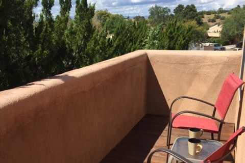 A private balcony features a wooden floor and a stucco wall, framed by greenery. Two red chairs are positioned around a small table, which holds a coffee cup, inviting guests to enjoy a peaceful outdoor space with views of the surroundings.