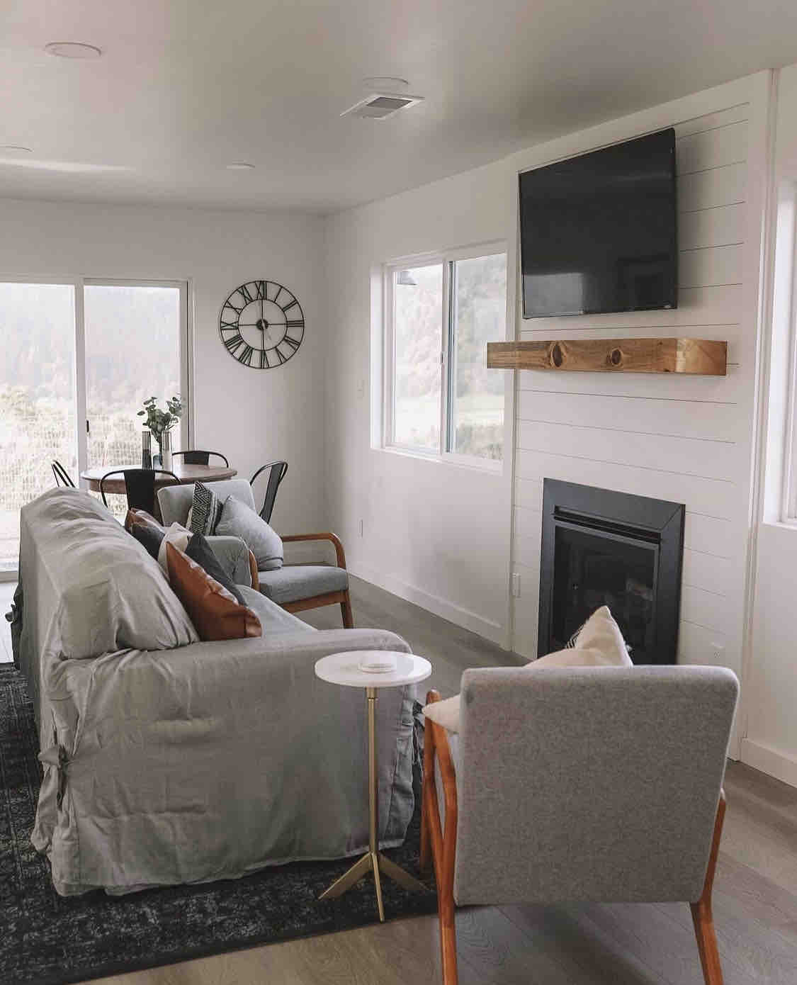 A cozy living area is furnished with a light grey sofa and comfortable chairs. A wooden shelf holds a flat-screen TV above a modern fireplace. Large windows allow natural light to fill the space, offering views of the surrounding landscape.