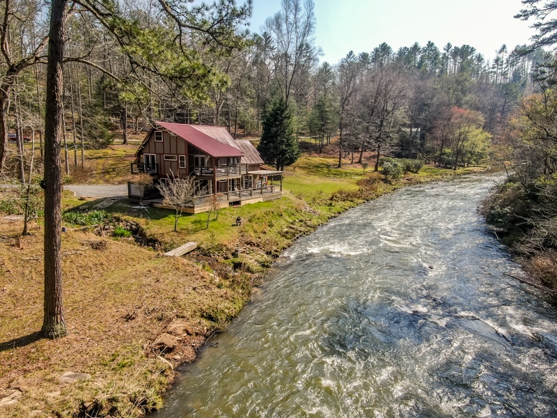 The rustic farmhouse is set alongside a flowing river, surrounded by lush greenery and trees. A spacious porch wraps around the front, inviting relaxation, while the water creates a calming sound as it gently moves past the property.