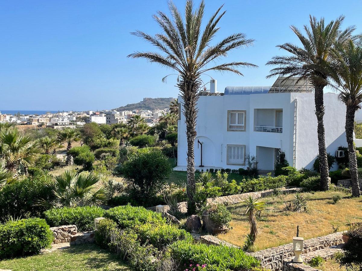 The image captures a modern white apartment surrounded by lush greenery and palm trees. The vibrant landscape leads towards a scenic view of the coastline in the distance. Clear blue skies enhance the sunlit atmosphere, providing a sense of tranquility and connection with nature.