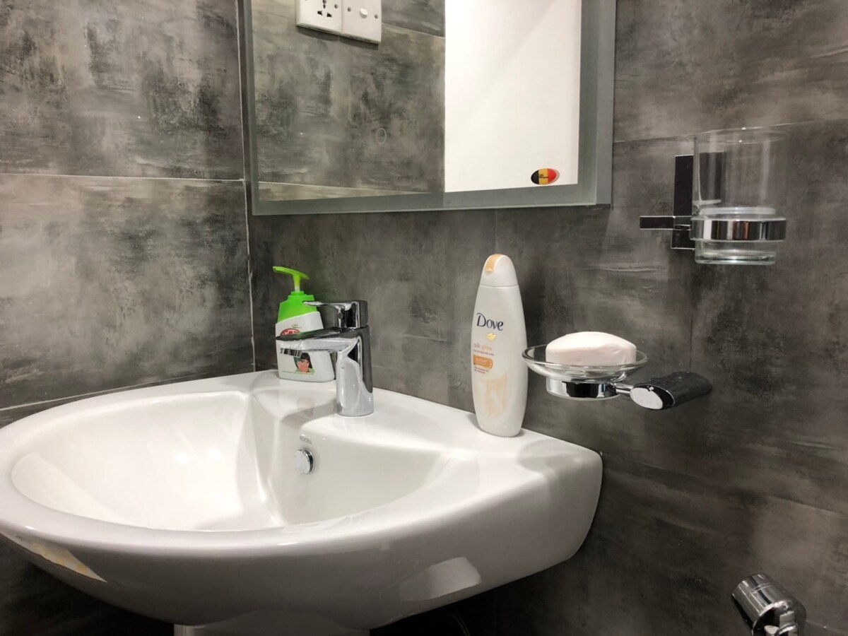 A modern bathroom sink area is displayed, featuring a sleek faucet and a circular mirror above. Soap and a bottle of body wash are neatly positioned beside the sink. A holder for a toothbrush and a small green bottle are visible, complemented by a glass for rinsing.