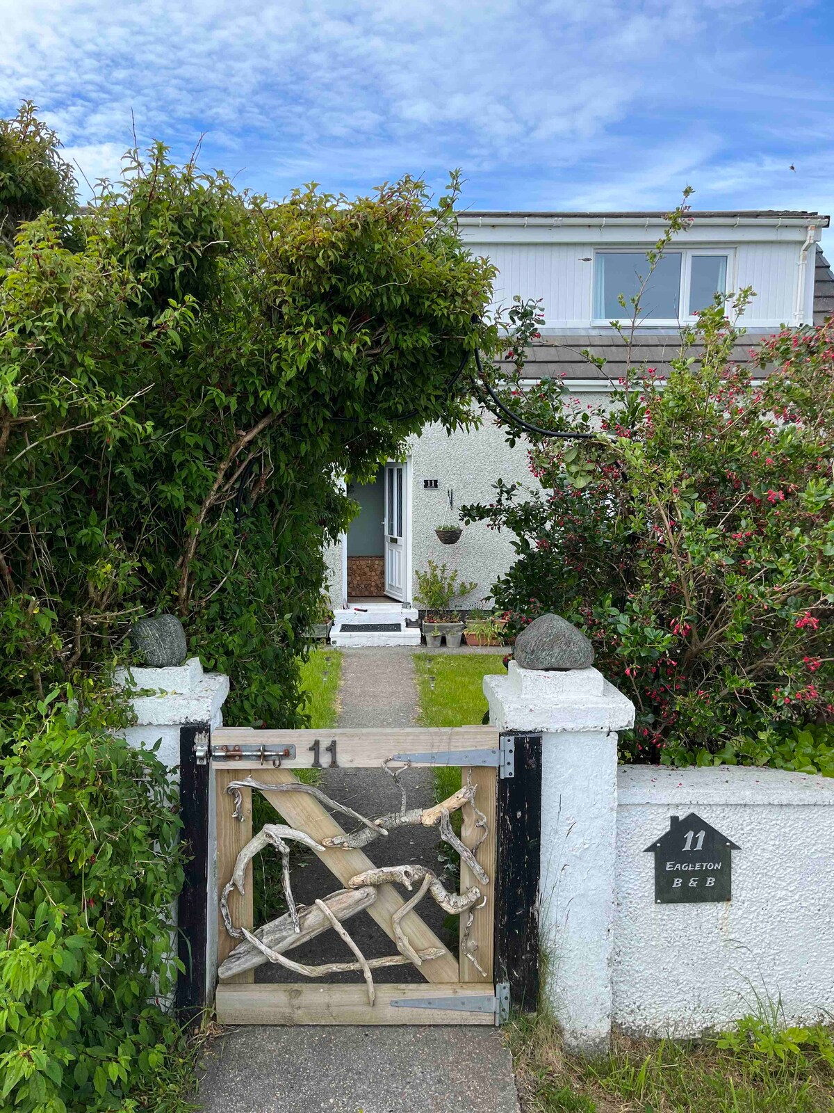 A modest exterior view of Eagleton B&B is framed by lush greenery and a wooden gate. The pathway leads to the inviting entrance, which is adorned with simple decorative elements. A house number sign is visible on the white wall, indicating the location.