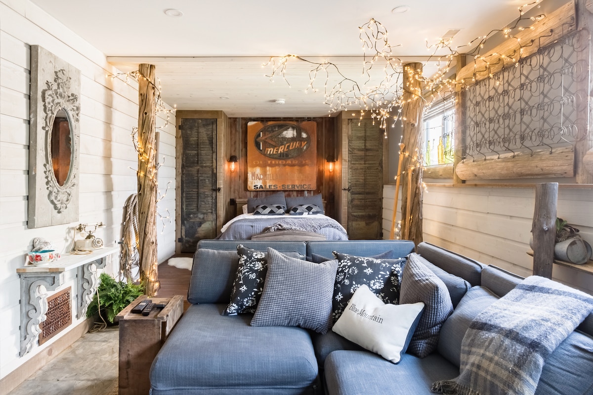 A cozy studio space is illuminated by soft string lights, complementing the rustic decor. An inviting seating area features plush cushions on a sectional sofa, while a spacious bed is set against a backdrop of reclaimed wood. Vintage elements add character to the room.