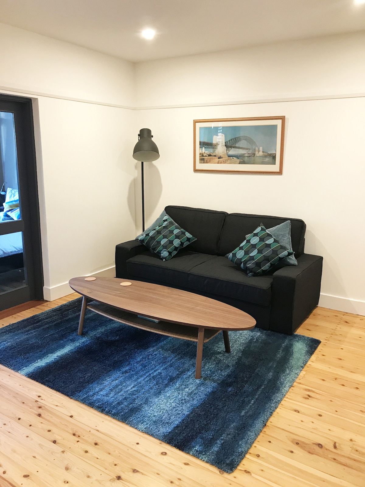 A cozy lounge area features a dark couch with decorative cushions resting on a vibrant blue rug. A sleek coffee table is positioned in front, and a floor lamp stands beside the couch. Artwork depicting a harbor scene adorns the wall.