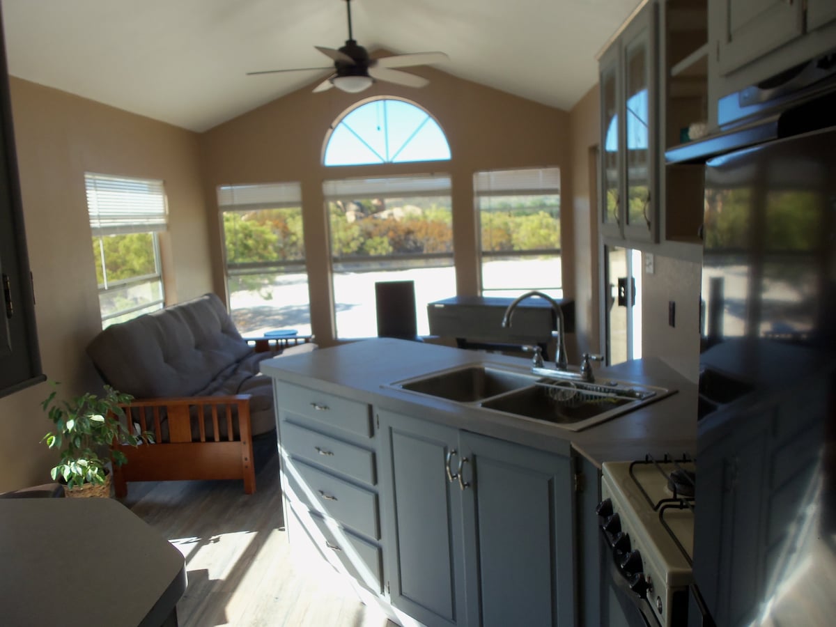A light-filled interior features a modern kitchen with gray cabinetry and a double sink. A comfortable sitting area includes a convertible sofa, with a large window providing views of the surrounding landscape. Sunlight streams through the windows, enhancing the space's openness.
