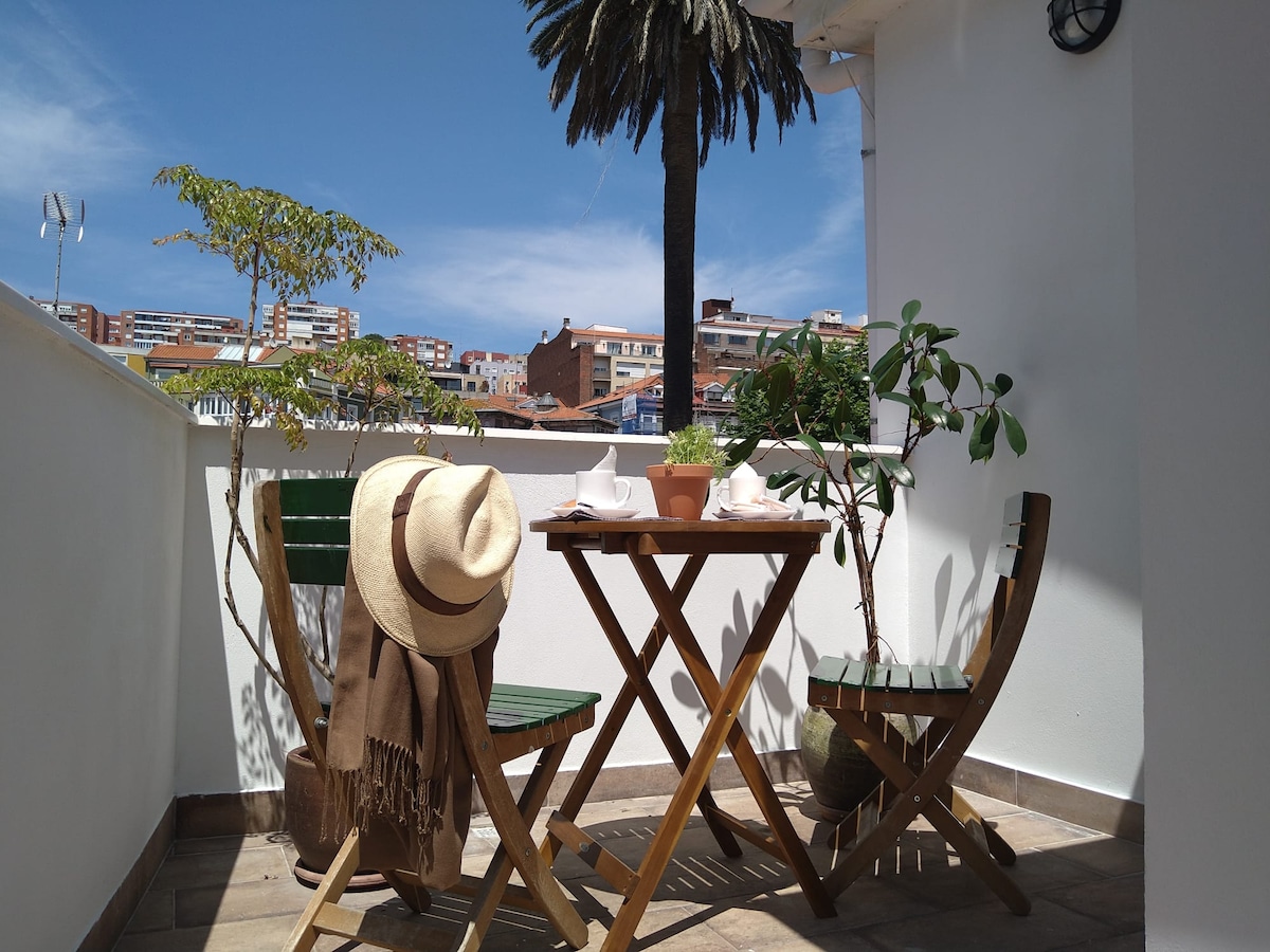 A small terrace features a wooden table surrounded by two folding chairs. Potted plants add greenery, and a palm tree stands in the background. A straw hat and brown shawl rest on one chair, inviting relaxation in the sunlight.