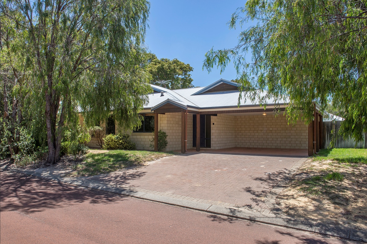A family home is situated on a quiet street, framed by trees and foliage. The exterior features a single-story structure with a mix of brick and weatherboard finishes, providing a welcoming entrance along the pathway leading to the front door.