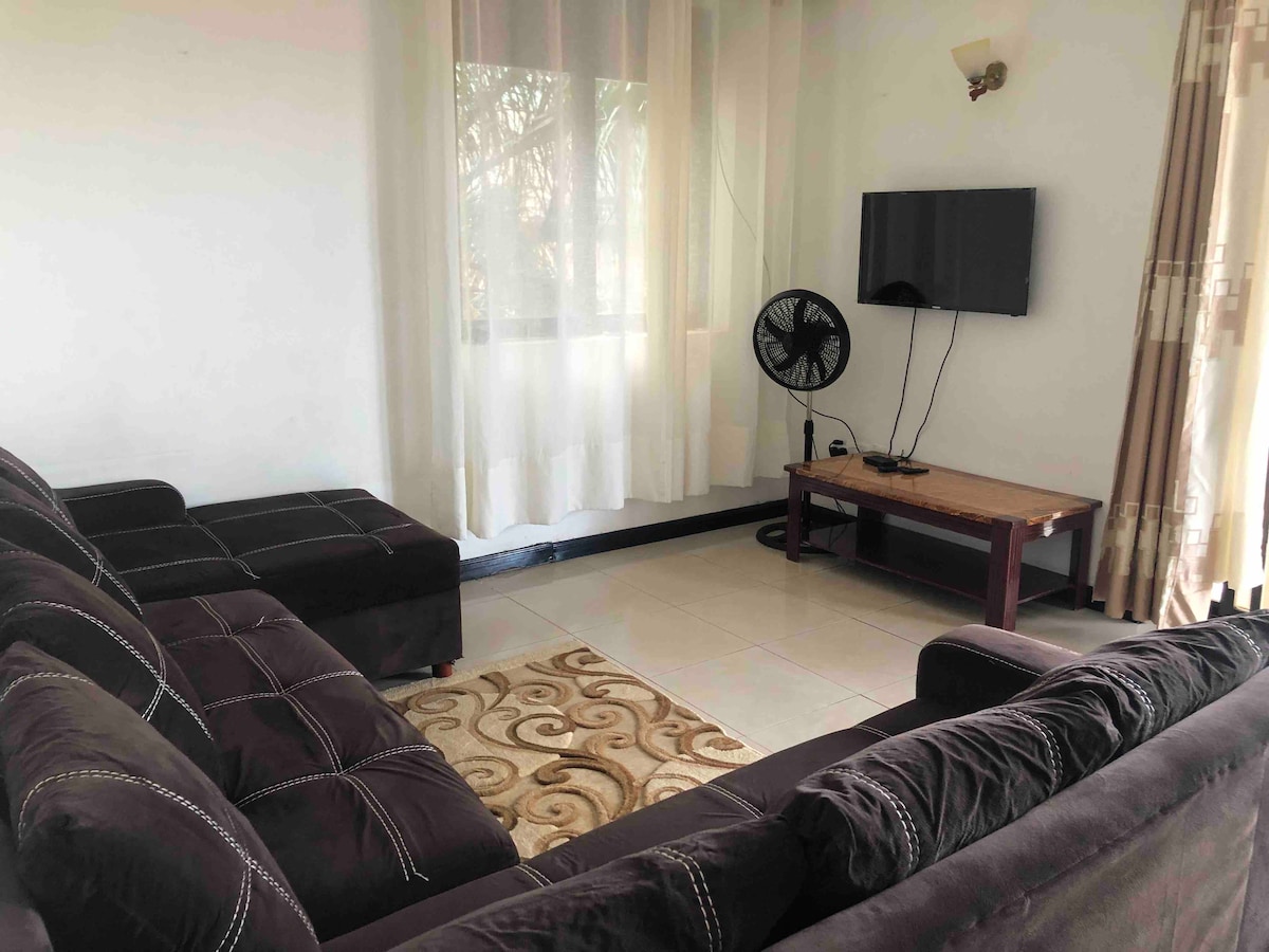 A comfortable living area includes a dark upholstered sectional sofa, a wooden coffee table, and a flat-screen TV mounted on the wall. Natural light filters through sheer white curtains, enhancing the space's openness. A floor fan is positioned nearby for added comfort.