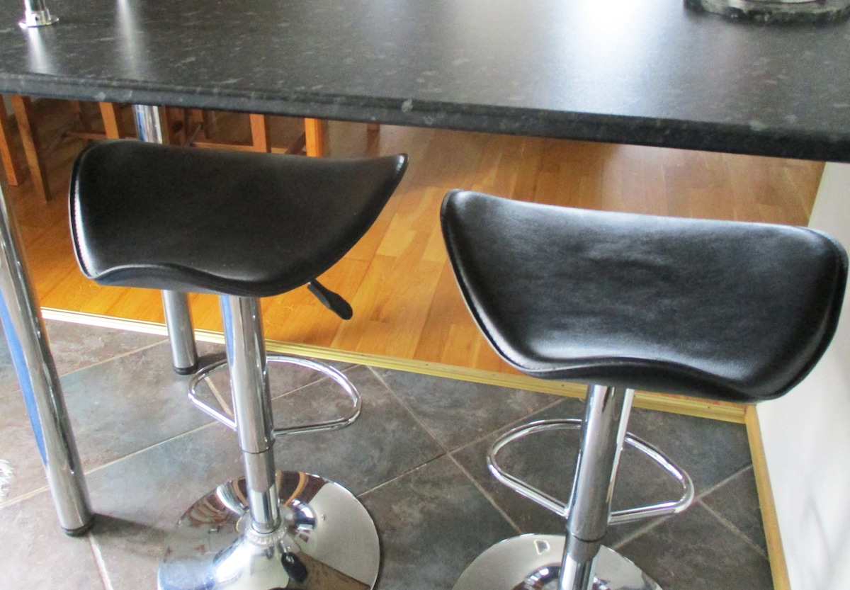 Two black bar stools with a sleek design are positioned beneath a dark, polished countertop. The shiny, reflective bases contribute to a modern look. The floor features a combination of tiles and wooden elements, providing a balanced contrast.