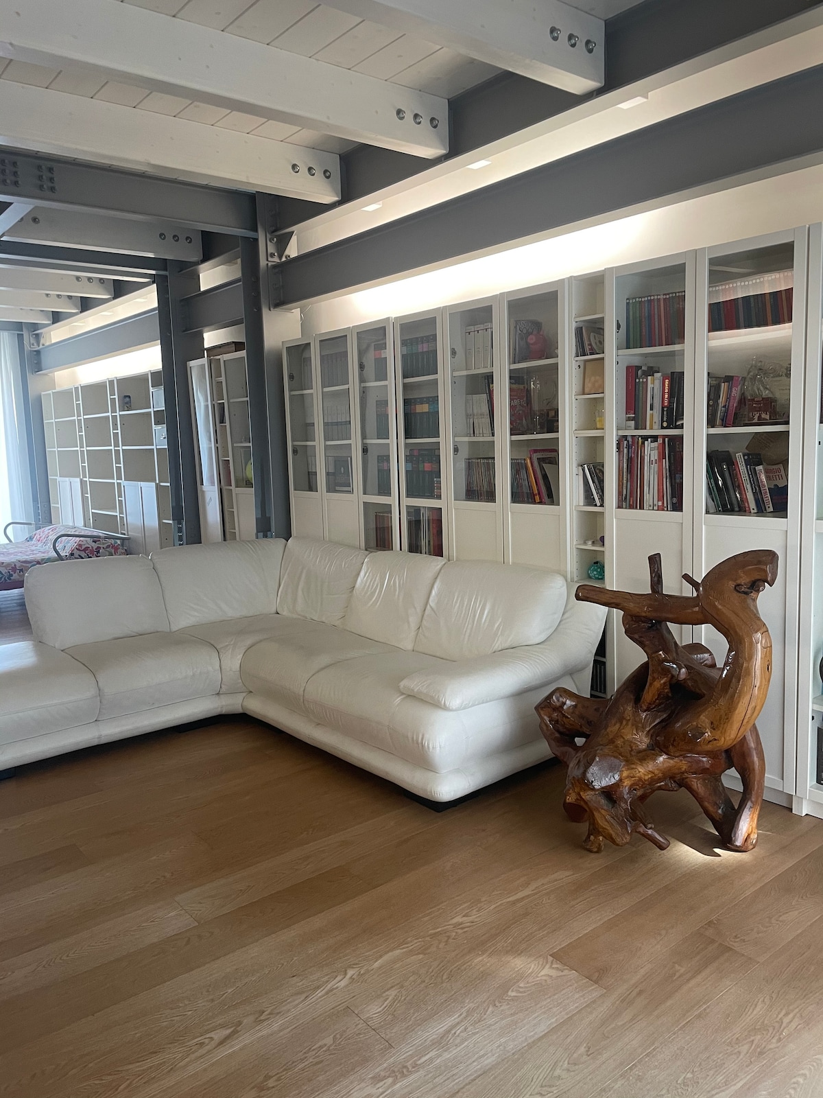 A white sectional sofa is positioned near a wall of glass-fronted bookshelves filled with various books. The warm wooden flooring complements the artistic wooden sculpture displayed nearby, contributing to a modern yet inviting living space.