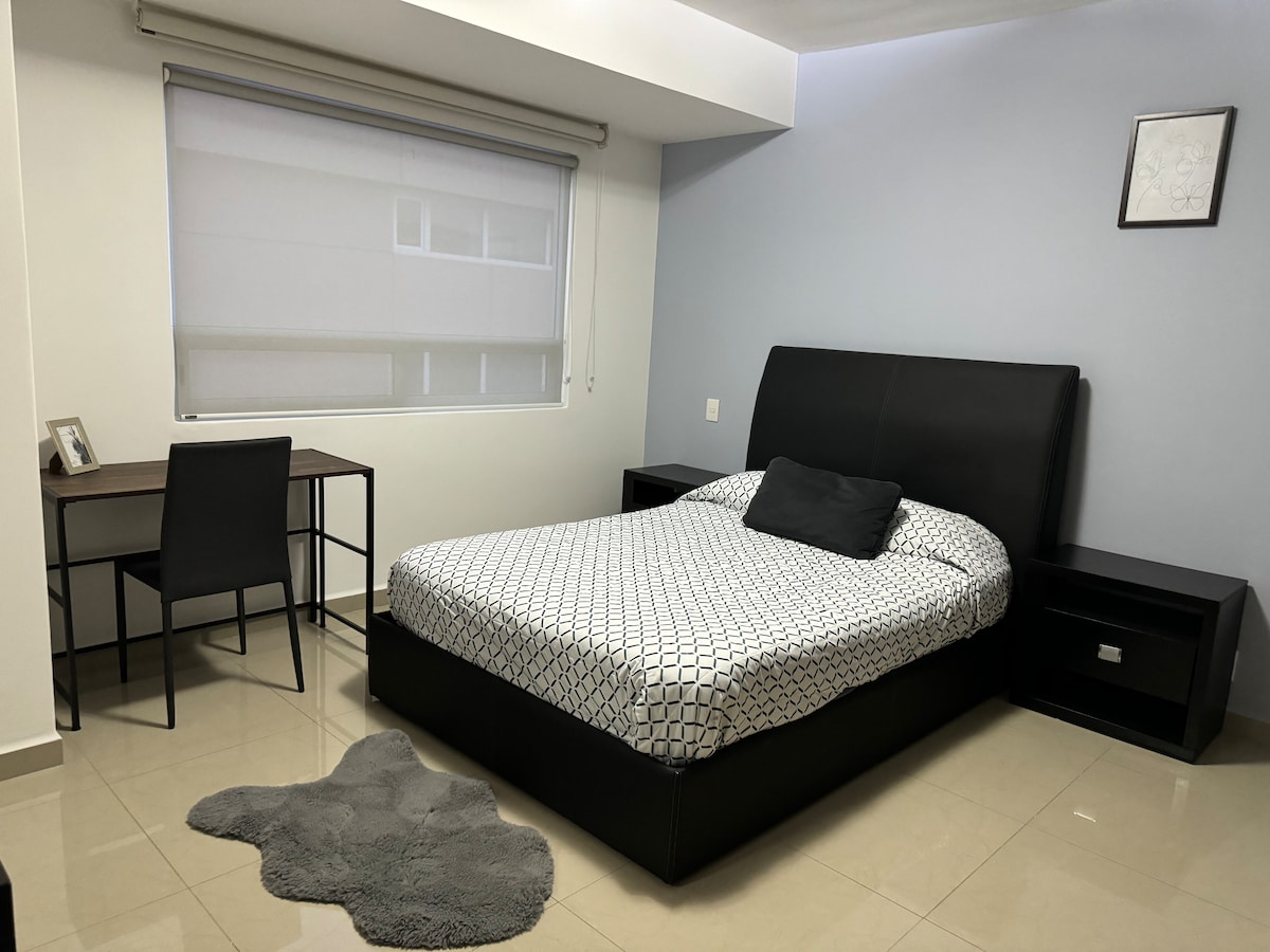 A well-lit bedroom is presented, featuring a spacious bed with a textured duvet and a dark headboard. A small desk with a chair is positioned next to the bed. A window with light-filtering shades allows natural light to enter, enhancing the calming ambiance.