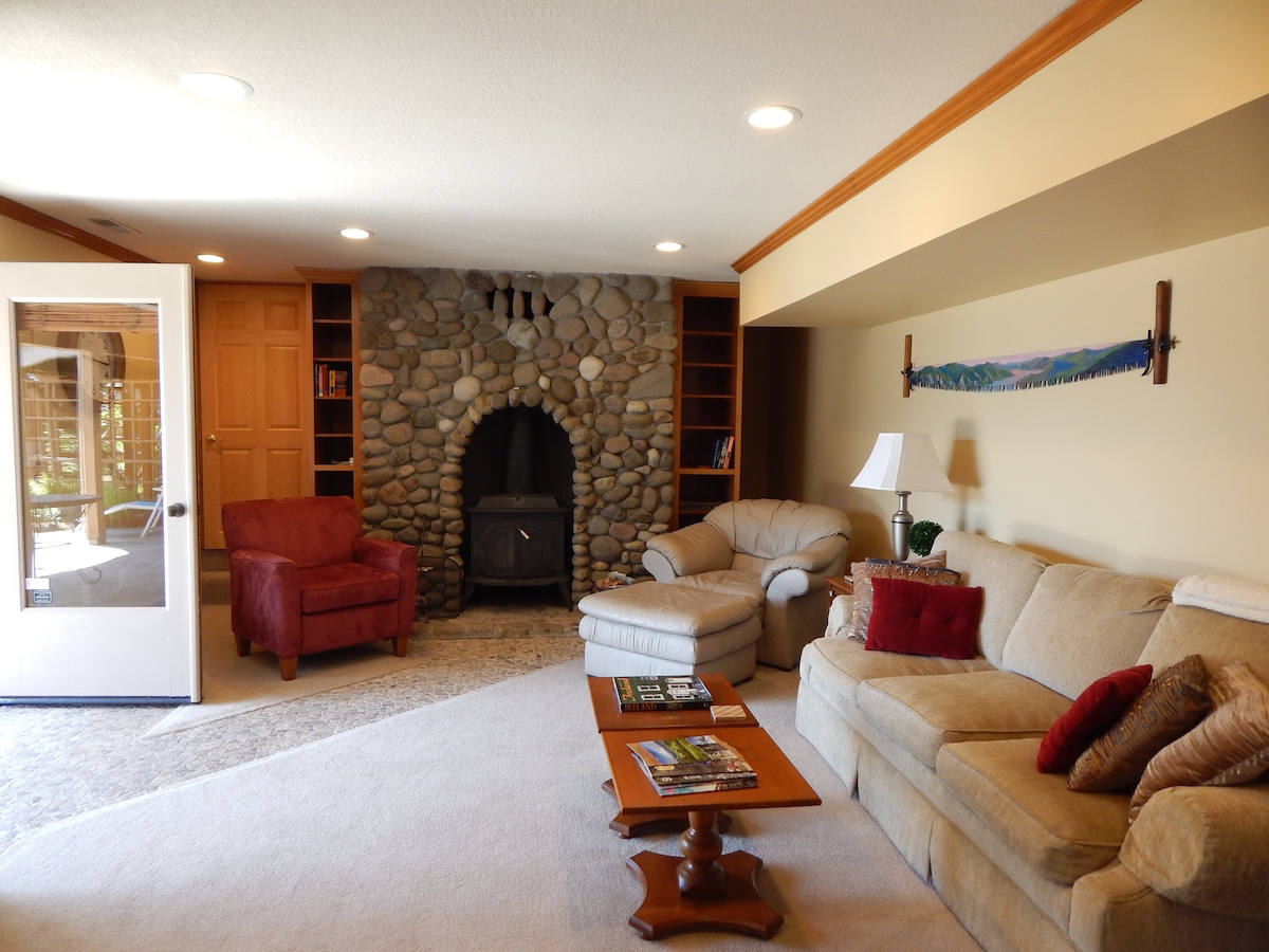 A cozy living room is depicted, featuring a river rock fireplace as a focal point. Comfortable seating options include a beige couch and an armchair. A wooden coffee table holds stacked magazines, while built-in shelves display books. Natural light streams through the French doors.