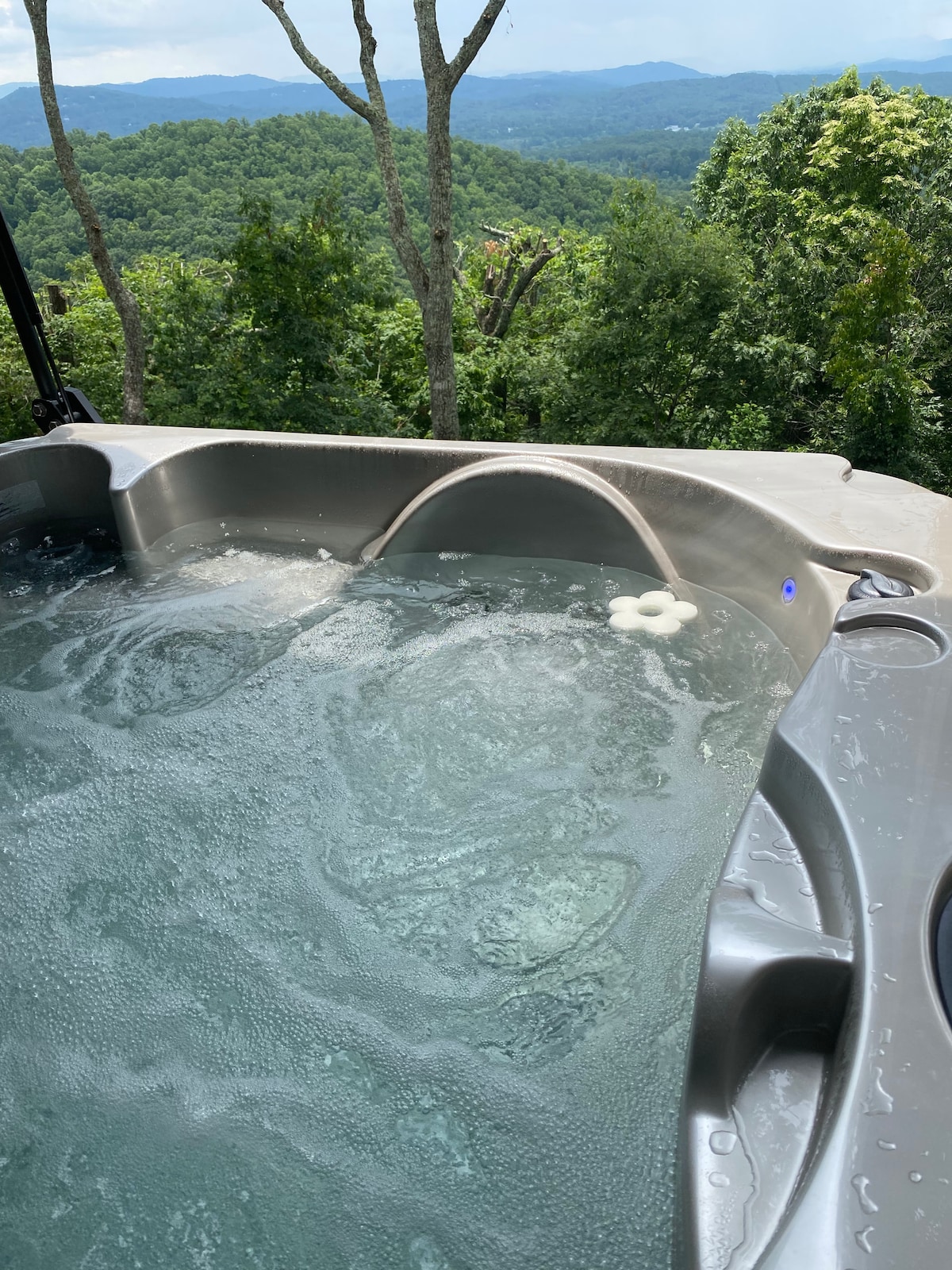 A two-person jacuzzi is filled with bubbling water, positioned on a deck surrounded by lush greenery. The scenic views of the treetops and distant mountains are visible in the background, enhancing the tranquil atmosphere.