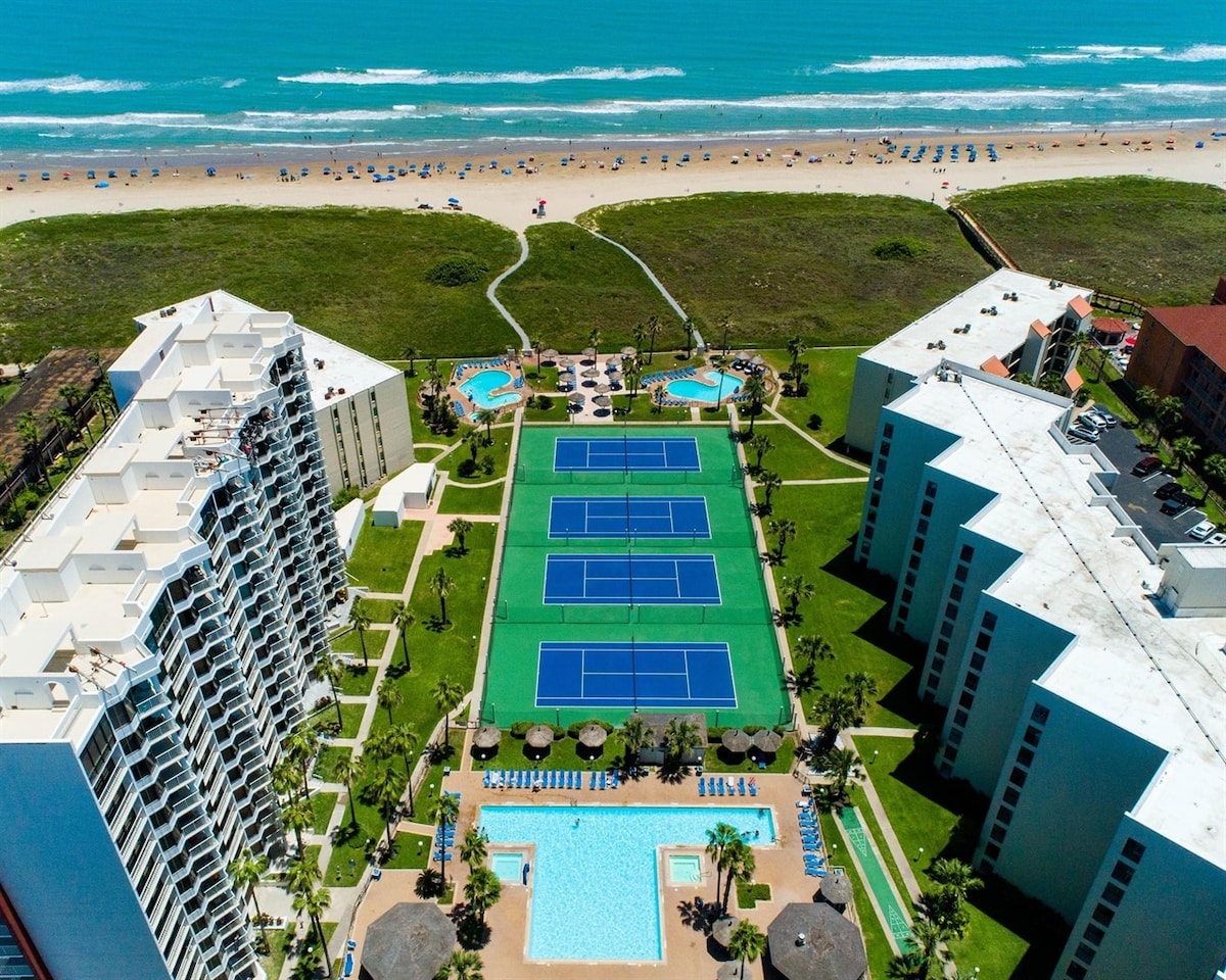 An aerial view displays the resort’s expansive grounds, featuring three tennis courts and several swimming pools surrounded by lush landscaping. The beach is visible in the background, with beach chairs dotted along the shoreline and the vibrant turquoise ocean stretching out beyond.