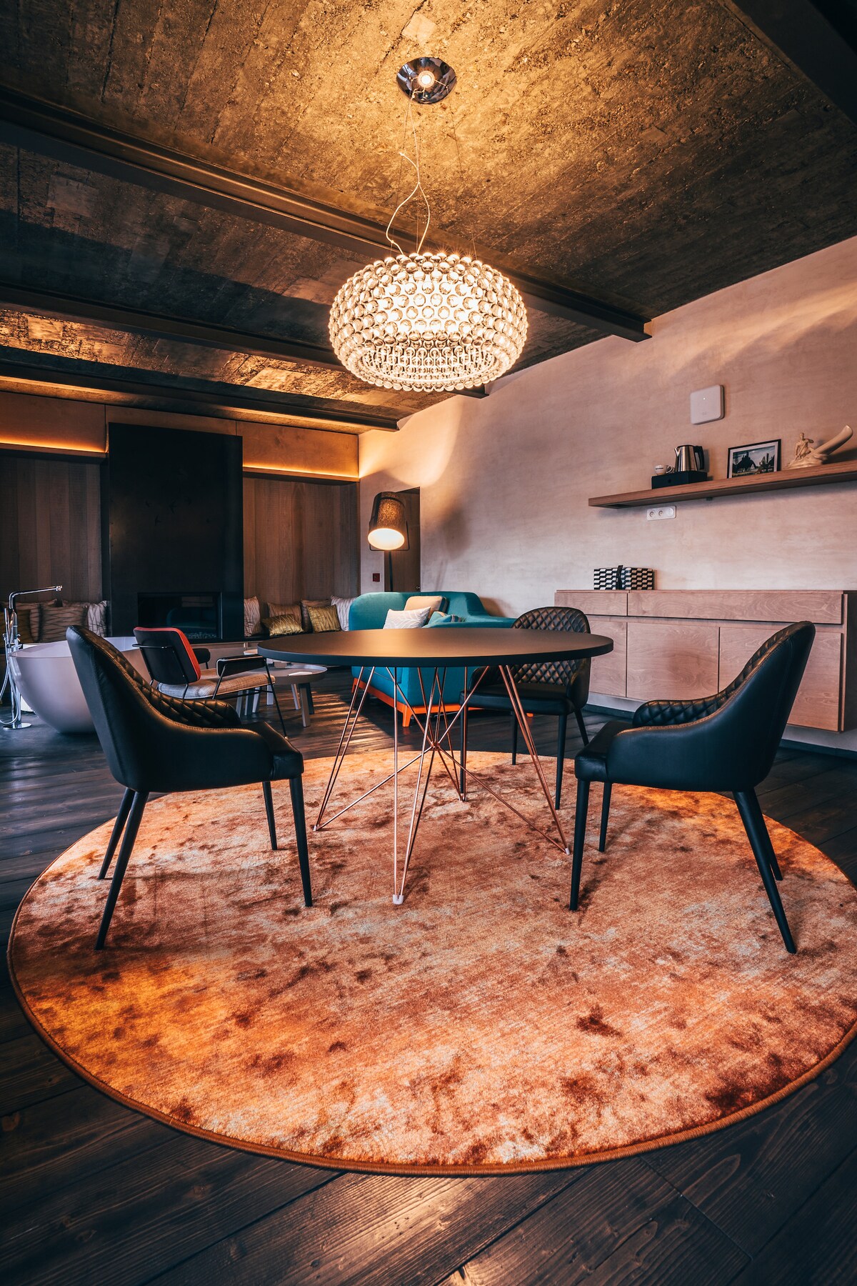 A comfortable sitting area is highlighted by a round area rug beneath a sleek dining table surrounded by modern black chairs. The space is softly illuminated by a decorative pendant light, with muted decor elements in the background creating a relaxed ambiance.