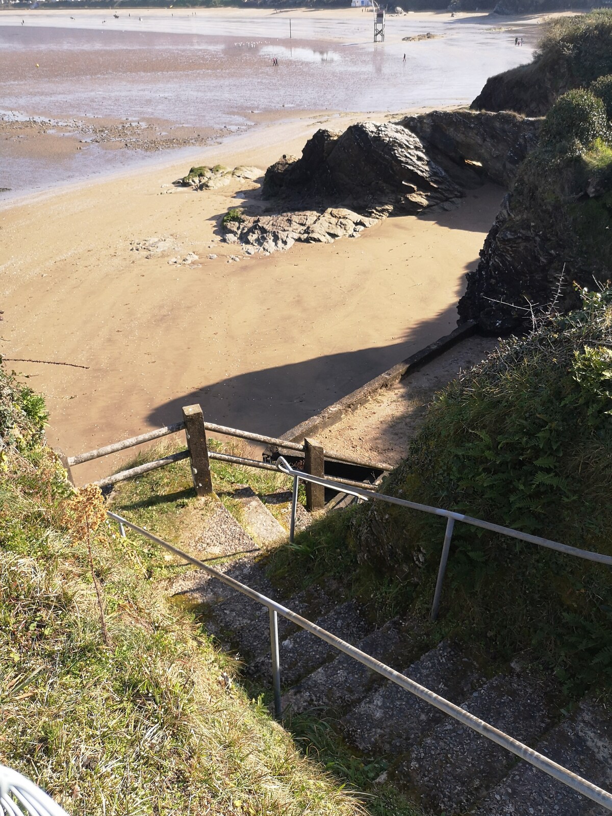 A descending staircase is viewed from the top, leading down to a sandy beach. Alongside, greenery frames the pathway, while rocky formations are visible on the beach. The shoreline extends into the background, with people seen enjoying the coastal area.