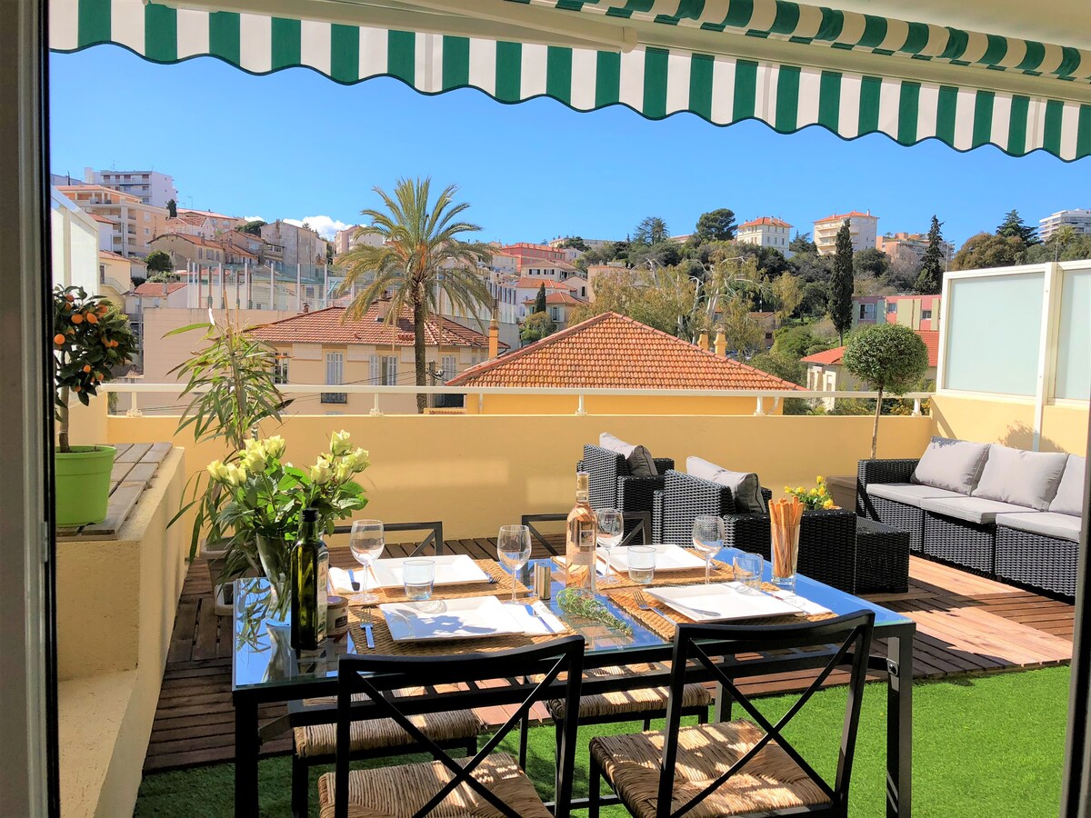 A spacious terrace features a dining table set for four, accompanied by comfortable seating areas. The area is adorned with greenery and offers a view of surrounding buildings and palm trees. Natural light brightens the space under a striped awning.