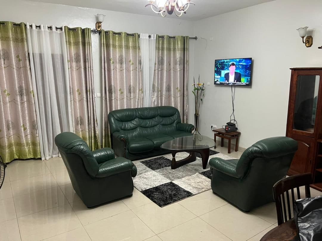 A spacious living area is presented with green leather sofas arranged around a round glass coffee table. A television mounted on the wall is visible, along with elegant curtain panels featuring a floral design. Light-colored tiles enhance the room's brightness.