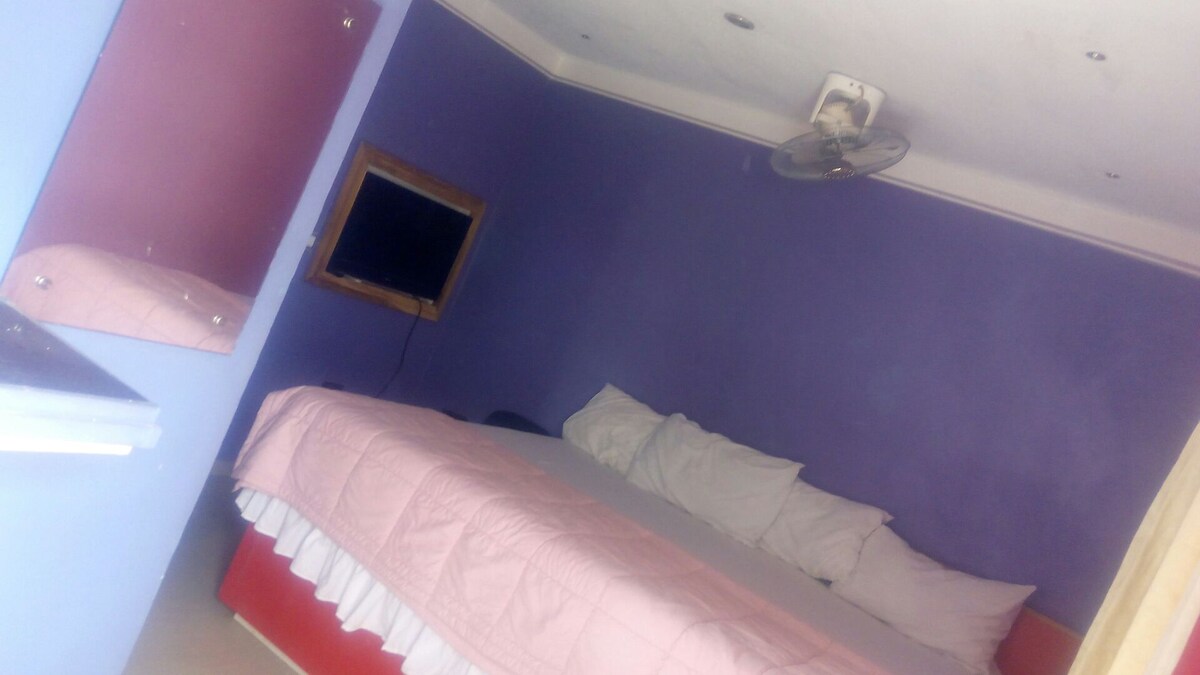 A sleeping area features a spacious bed adorned with pillows against a purple wall. A flat-screen TV is mounted on the wall above a wooden frame, while a mirror reflects the room's layout. Light-colored bedding contrasts with the deeper wall hue.