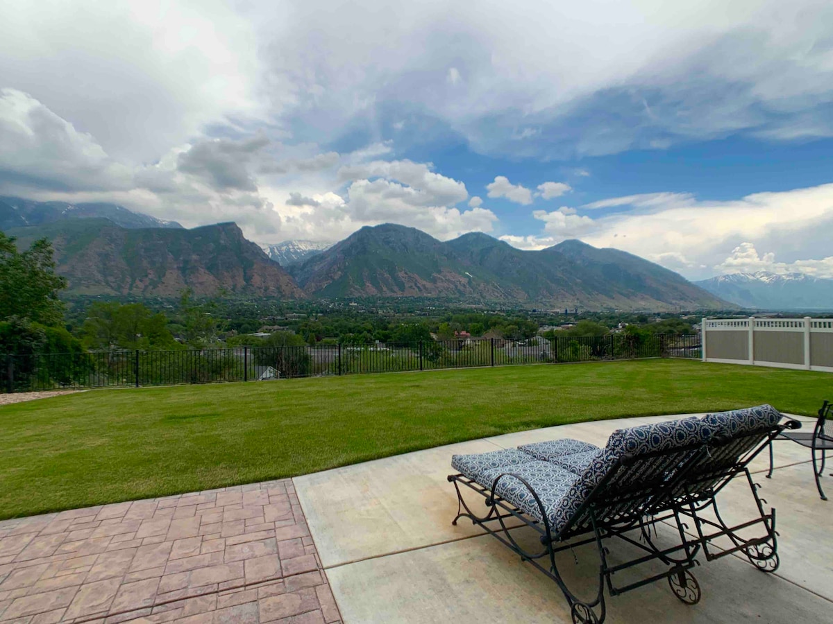 An outdoor space features a well-kept lawn with a view of distant mountains and cloudy skies. A lounge chair with a patterned cushion is positioned on the patio, overlooking the scenic landscape. A fence borders the property, enhancing the private atmosphere.