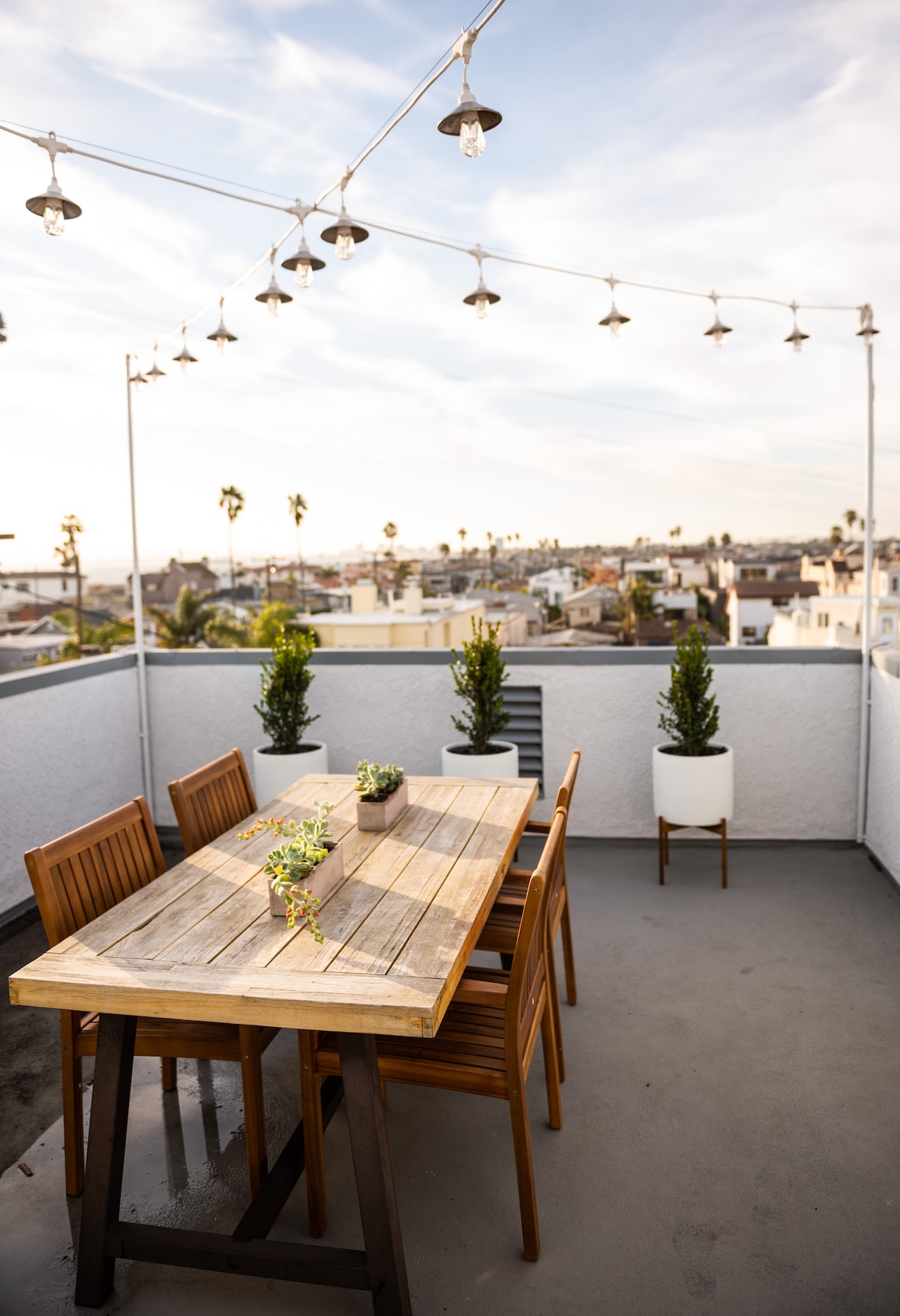 An outdoor dining area is showcased on the roof deck, featuring a wooden table surrounded by several chairs. Potted plants are placed strategically at the corners, and string lights hang overhead against a backdrop of the skyline and palm trees.