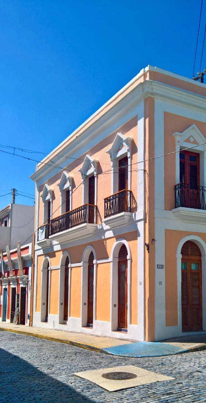 A historic two-story building is situated on a cobblestone street, featuring a light peach facade with arched windows and decorative elements. A small balcony is positioned above the entrance, enhancing the Spanish colonial architectural style that defines the charm of Old San Juan.