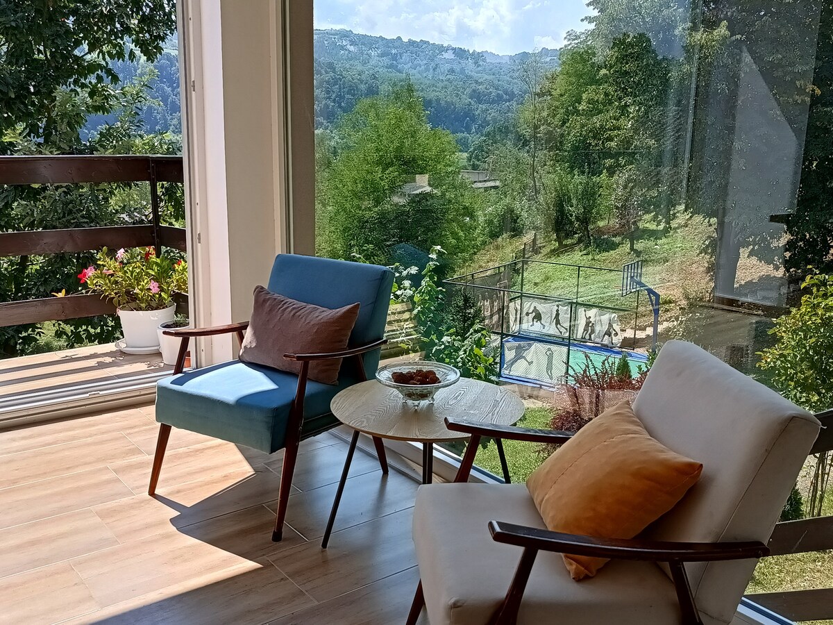 A cozy seating area features two chairs beside a small table, with natural light flooding through large windows. Outside, greenery and a fenced area are visible, suggesting a vibrant outdoor space ideal for relaxation and play.