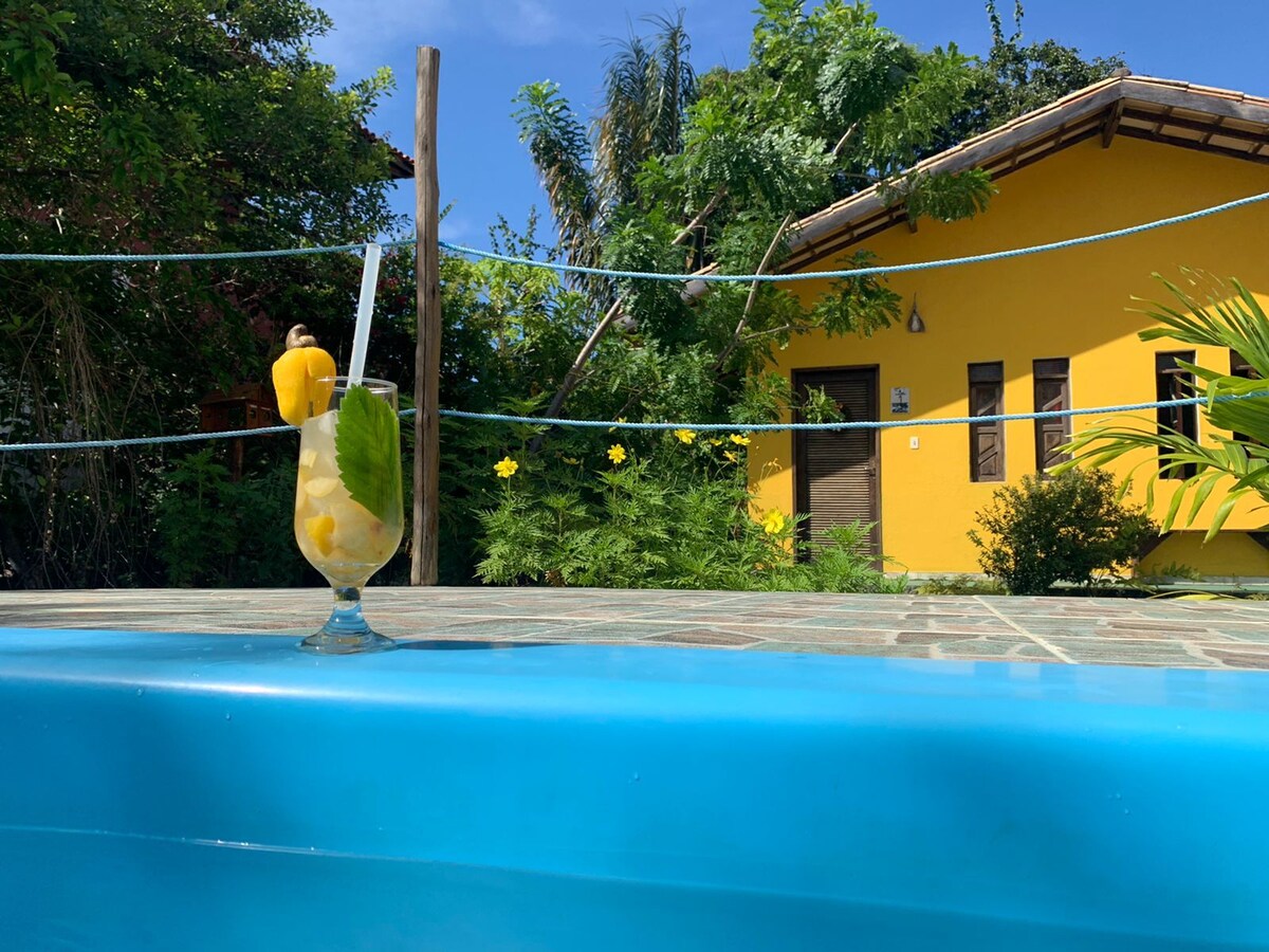 A refreshing beverage is placed on the edge of a swimming pool, with a bright yellow house visible in the background. Lush greenery surrounds the area, complementing the clear blue sky and inviting atmosphere.