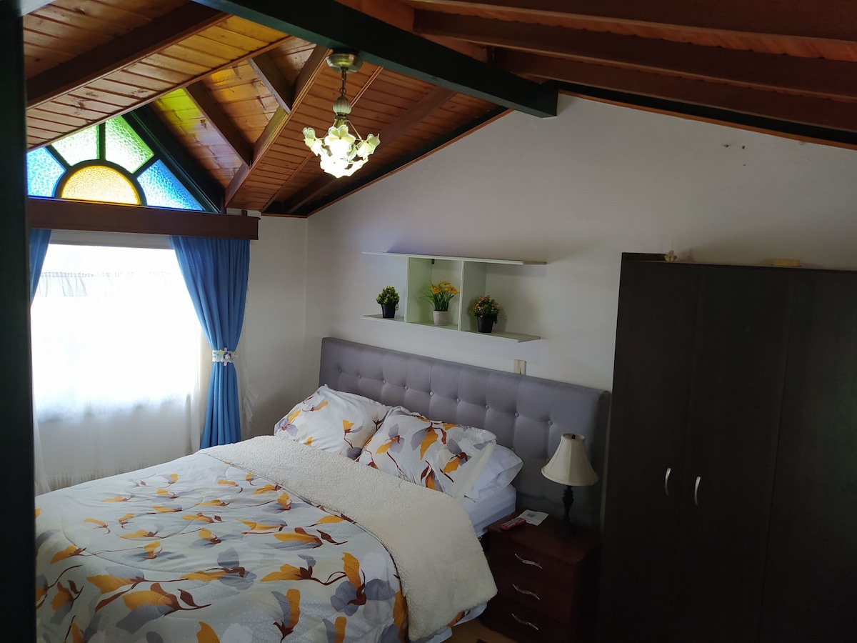 A cozy bedroom features a double bed with patterned bedding and decorative pillows. Natural light filters through a colorful stained glass window, enhancing the room's warmth. A nightstand and lamp are positioned beside the bed, while a closet stands against a wall adorned with decorative shelving.
