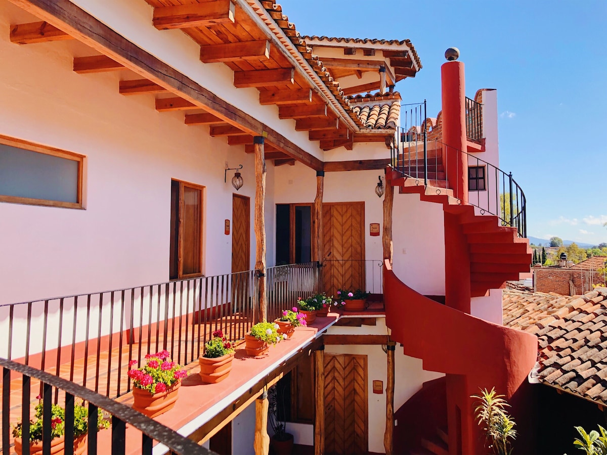 An inviting courtyard is framed by two levels of rooms, each featuring wooden doors and balconies with decorative railings. Vibrant flowers are arranged in pots along the walkways. A staircase spirals upward, connecting the levels against a backdrop of clear blue skies.