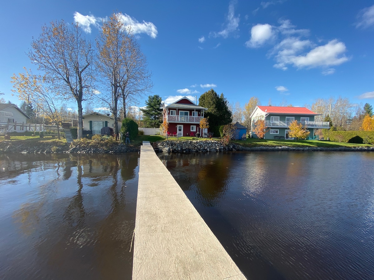 A solid dock stretches across calm waters, leading towards a charming two-story chalet surrounded by trees. The chalet features a balcony with a red roof, while the neighboring houses provide a backdrop of varied architectural styles against a clear blue sky.