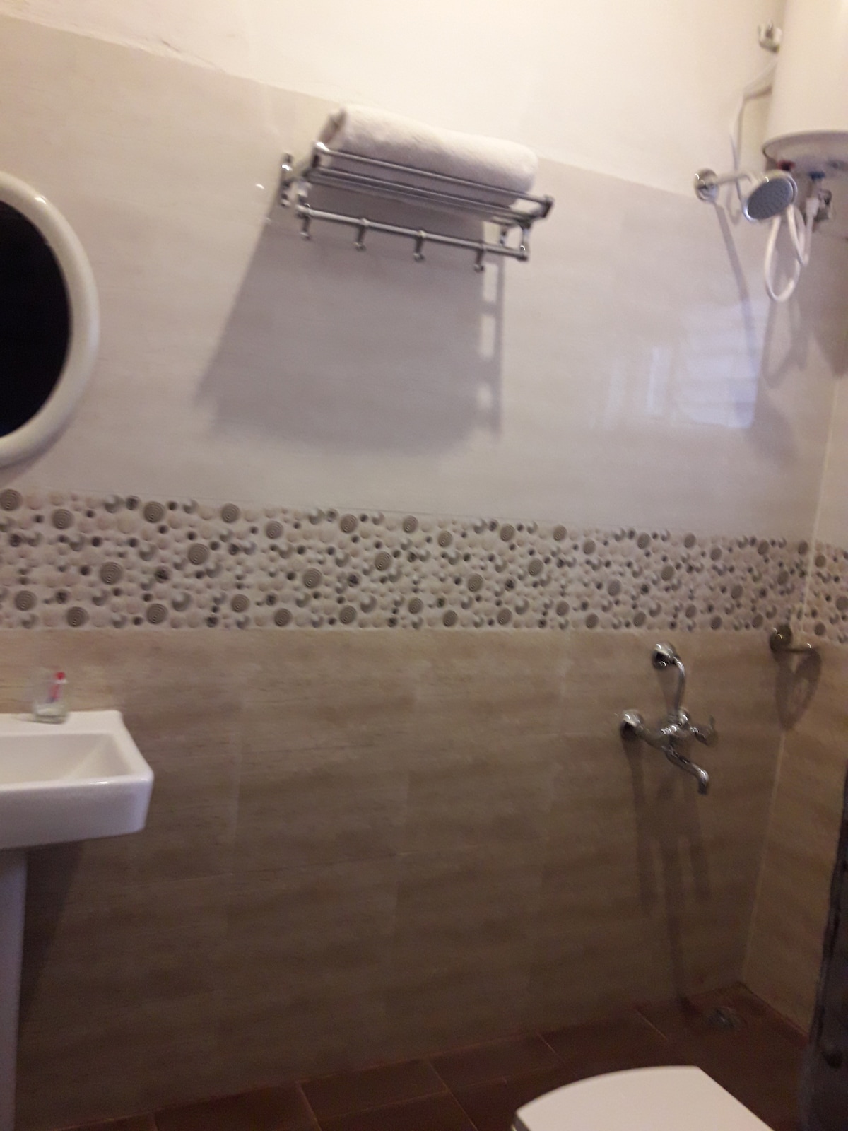 A clean bathroom space is presented, featuring a circular mirror above a modern sink. The walls are adorned with a decorative stone tile border, while a shower area is equipped with overhead and hand-held fixtures. A towel rack is positioned above for convenience.