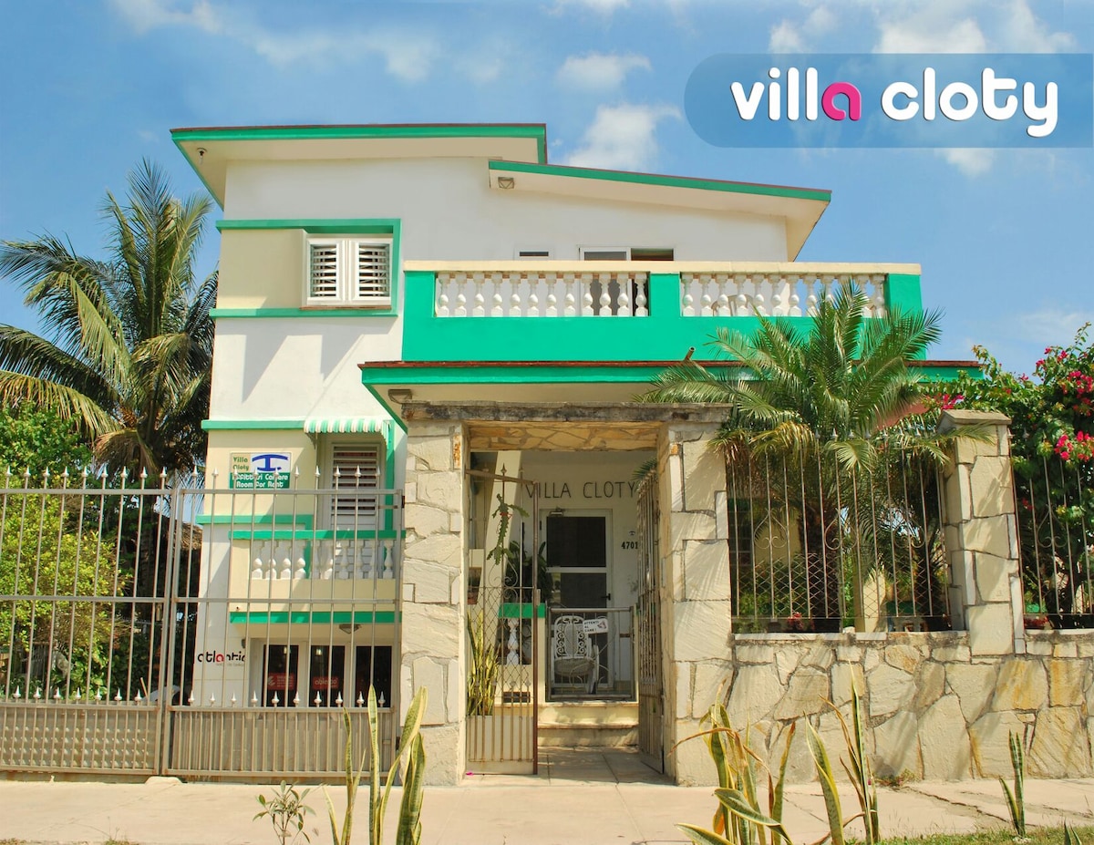 The exterior of Villa Cloty is presented, showcasing a two-story building with vibrant green accents. A gated entrance leads into the property, surrounded by tropical plants and palm trees. Clear blue skies and greenery are visible, enhancing the welcoming atmosphere of the villa.