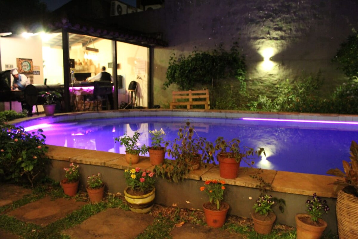An outdoor pool is illuminated with a soft purple glow, surrounded by potted flowers in vibrant colors. The background features a transparent wall that reveals a cozy indoor space, where guests can be seen enjoying time together.