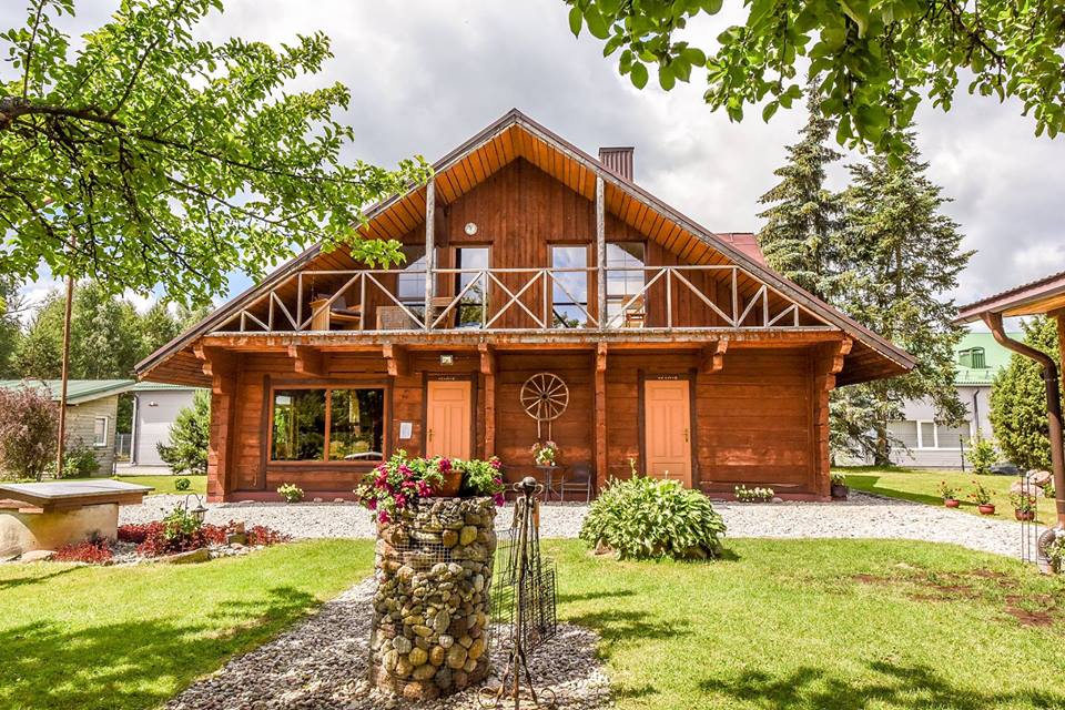 A two-story log cabin, crafted from Siberian wood, is showcased with large windows and a welcoming porch. Lush green lawns and colorful flowerbeds surround the cabin, while trees provide shade, enhancing the serene rural atmosphere.