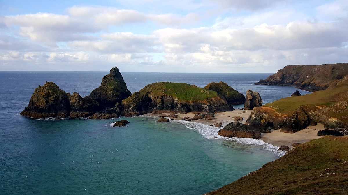 A coastal view showcases rugged cliffs and green hills meeting a calm sea. Several rocky formations emerge from the turquoise water, while a sandy beach is visible in the foreground. The sky is partly cloudy, adding depth to the serene landscape.
