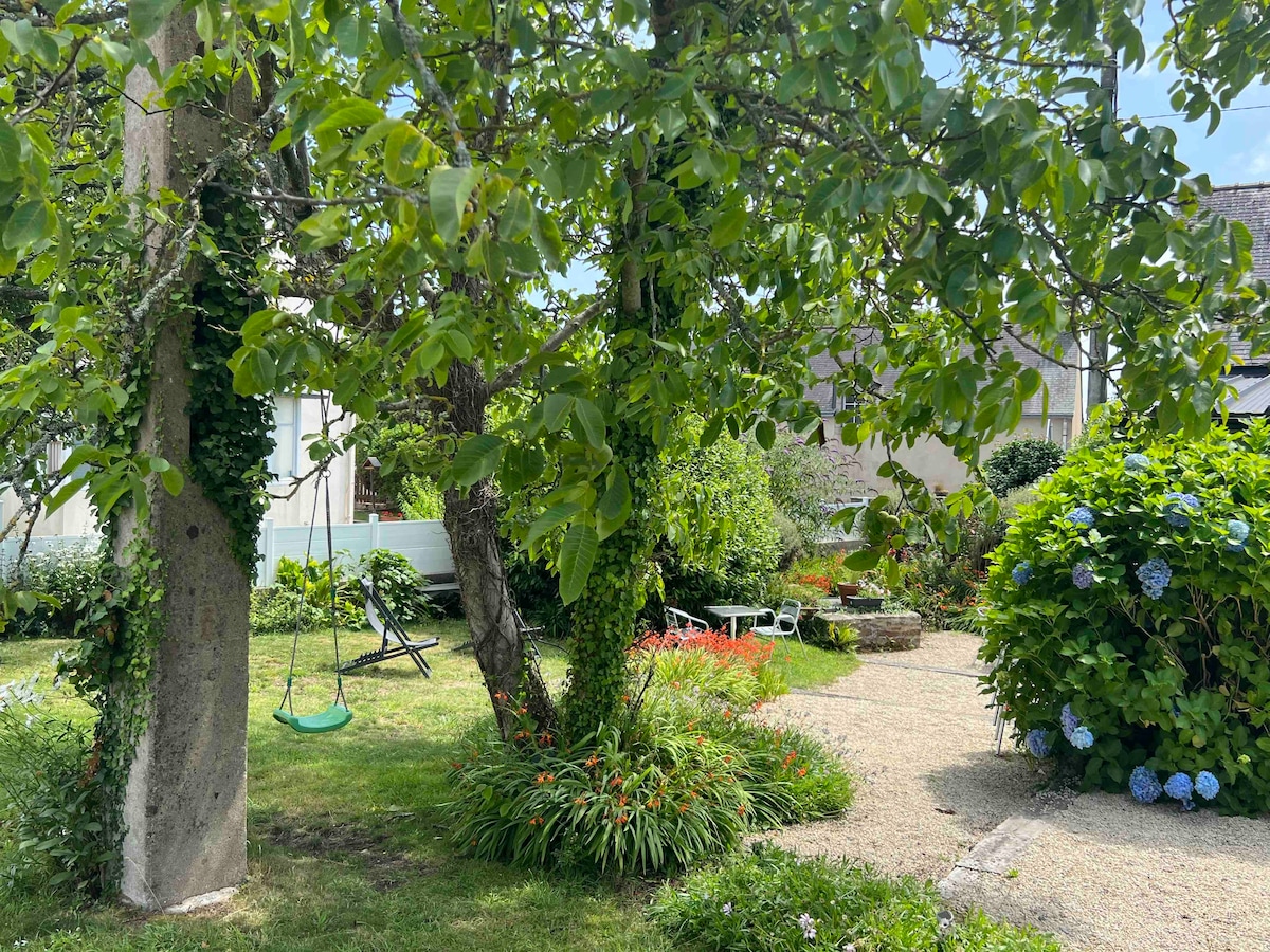 The garden features a lush landscape with vibrant greenery and blooming flowers. A swing hangs from a tree, inviting relaxation, while a stone path leads through the well-maintained space. Various seating areas are positioned throughout, offering options for enjoying the outdoors.