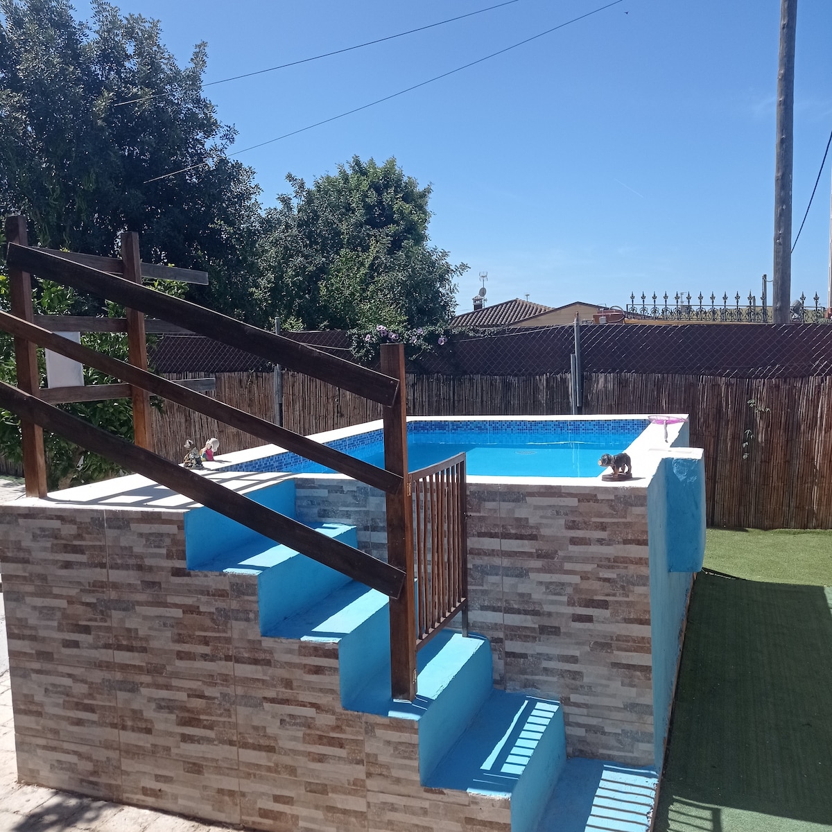 A private swimming pool features a blue tiled interior, surrounded by a stone wall. Steps leading up to the pool area are framed by wooden railings. Lush greenery fills the background, providing a serene outdoor setting.