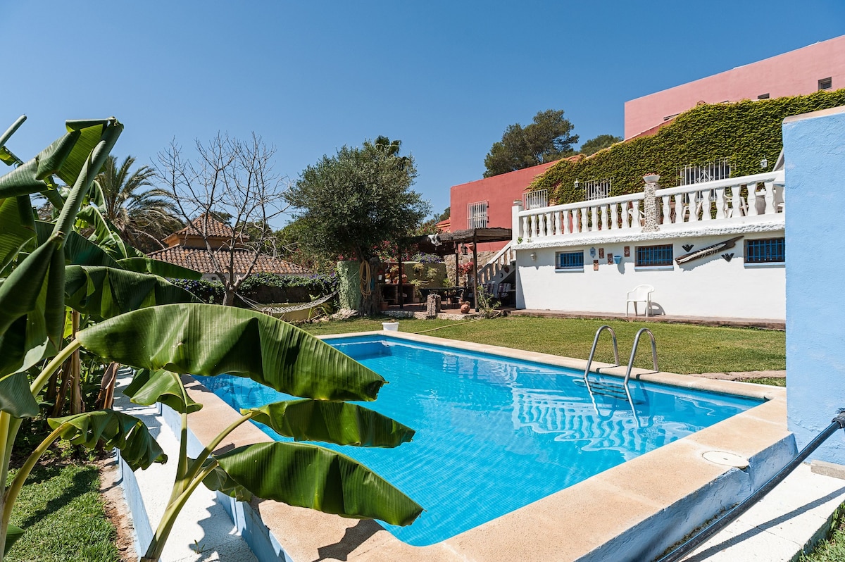 A well-maintained outdoor space features a clear blue swimming pool surrounded by lush greenery and palm trees. A staircase leads to a raised terrace with a view of the property, which displays elements of traditional architecture and colorful exterior walls.