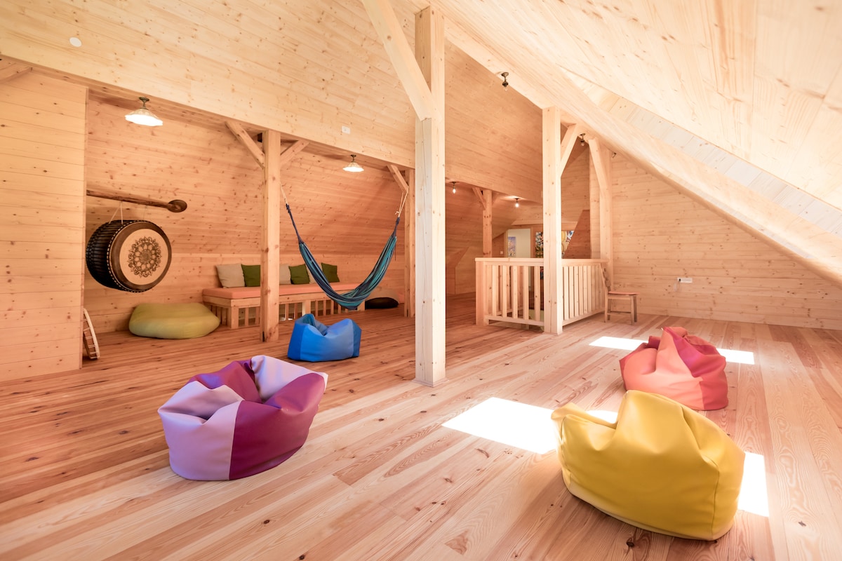 An airy loft space showcases wooden beams and a light-coloured wooden floor. Several colourful bean bags are arranged on the floor along with a hammock and a wooden table with chairs. Natural light floods the area from the sloped ceiling windows.