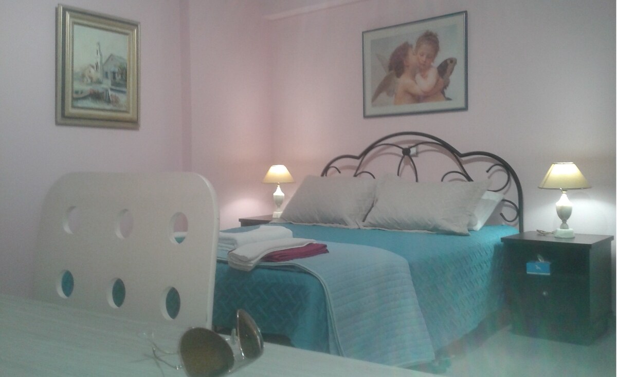 A cozy studio space features a spacious bed adorned with light-colored linens and a vibrant turquoise blanket. Soft lighting is provided by bedside lamps on each side of the bed, while a framed painting adds an artistic touch to the pink walls. A white table is visible in the foreground.