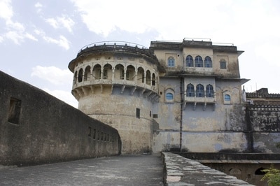 The historical structure features a circular turret alongside a rectangular main building with multiple large windows. The exterior exhibits weathered stone walls and decorative elements, with a pathway leading to the entrance, enhancing the fort's rustic charm.