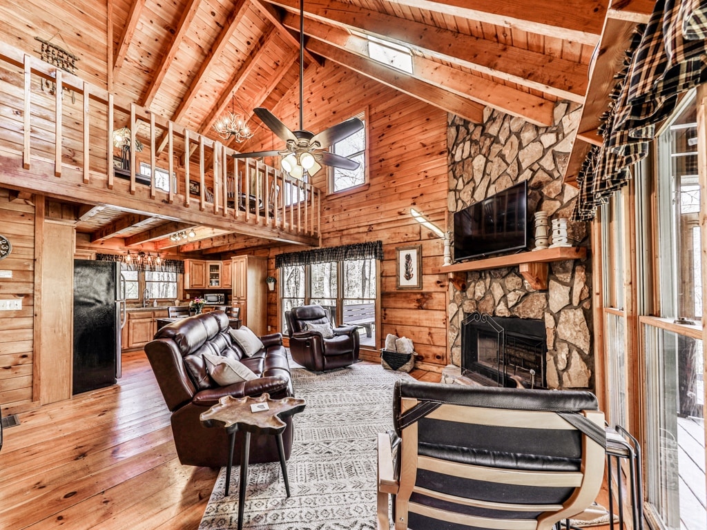 A cozy living area features wooden walls and a high ceiling with exposed beams. Two leather recliners are positioned around a coffee table, adjacent to a stone fireplace. A large flat-screen TV is mounted above the fireplace, with ample natural light entering through multiple windows.