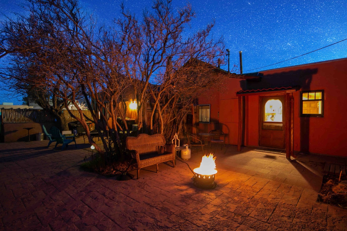 An outdoor space is seen, featuring a cozy seating area with wooden chairs and a small fire pit. Warm flames flicker within the pit, illuminating the surrounding trees against a starry night sky. A welcoming entrance can be seen in the background.